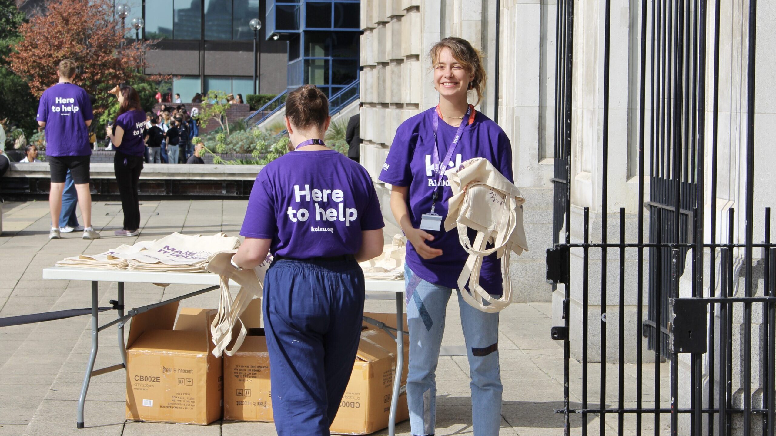 A photo of two KCLSU workers at King's Freshers' Fair. They are both wearing purple t-shirts, one has her back to the camera. Her t-shirt says "here to help". The other is smiling.