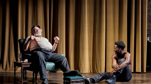 Ian McKellen as Falstaff sits on a chair on stage right. Toheeb Jimoh, as Prince Hal, looks at McKellen from his seat on the floor.