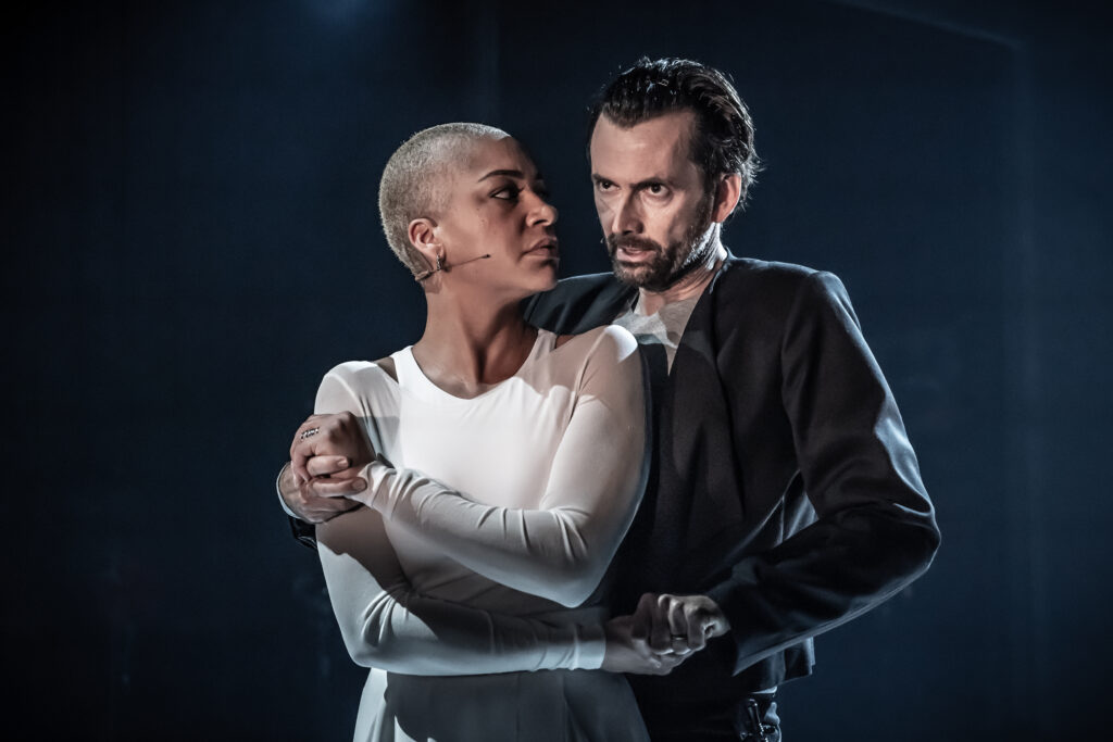 David Tennant as Macbeth holds Cush Jumbo as Lady Macbeth from behind. Tennant is dressed in black, and Jumbo in white.