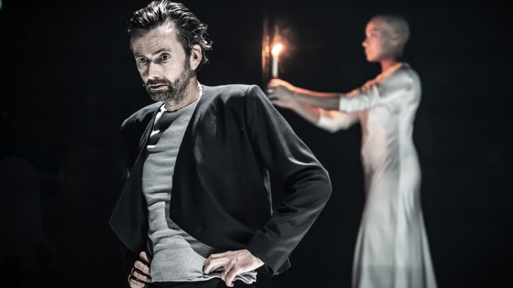 David Tennant as Macbeth stands in the foreground with his hands on his hips. In the background, Cush Jumbo as Lady Macbeth, in a white dress, carries a candle.