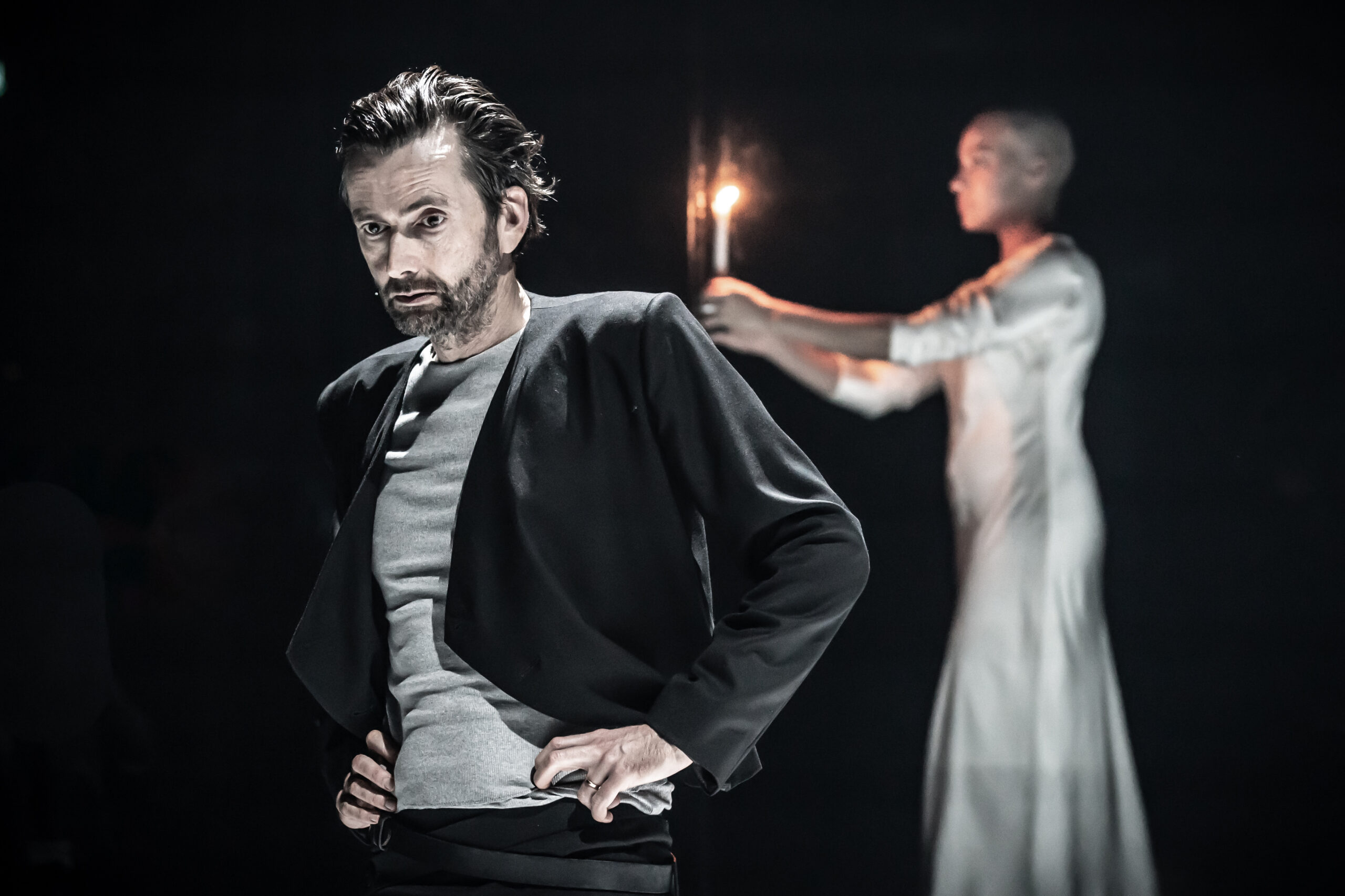 David Tennant as Macbeth stands in the foreground with his hands on his hips. In the background, Cush Jumbo as Lady Macbeth, in a white dress, carries a candle.