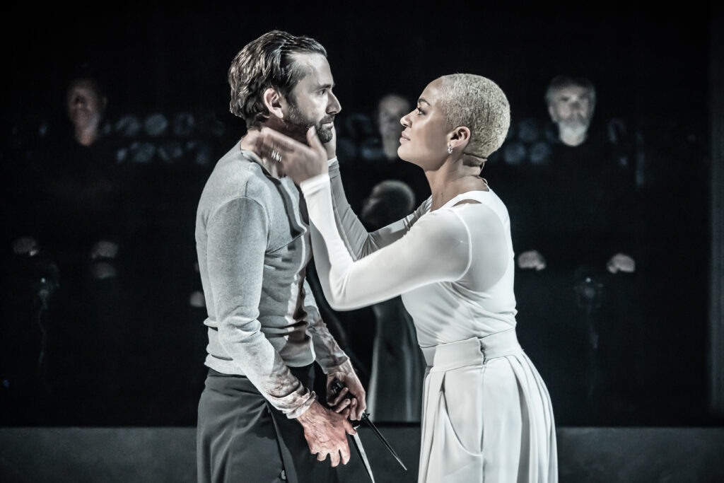 Cush Jumbo (Lady Macbeth) cups David Tennant's (Macbeth) face. They stare at each other—Tennant's arms are tense, and his face is grim.