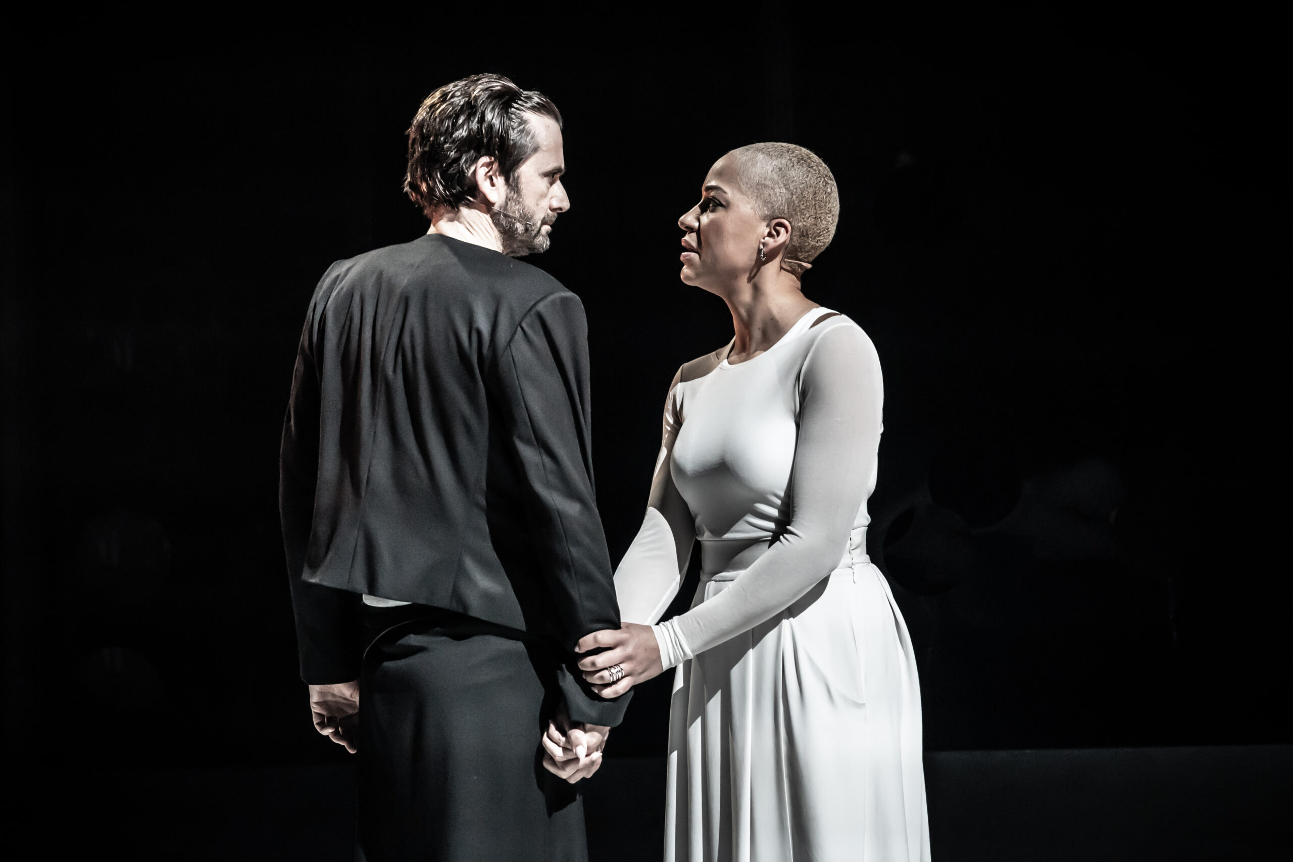 David Tennant, as Macbeth, faces Cush Jumbo, as Lady Macbeth. Tennant is in black, while Jumbo wears a white dress. Jumbo faces the camera, while Tennant's back is turned, and Jumbo holds Tennant's wrist. They're looking intently in each other's eyes.