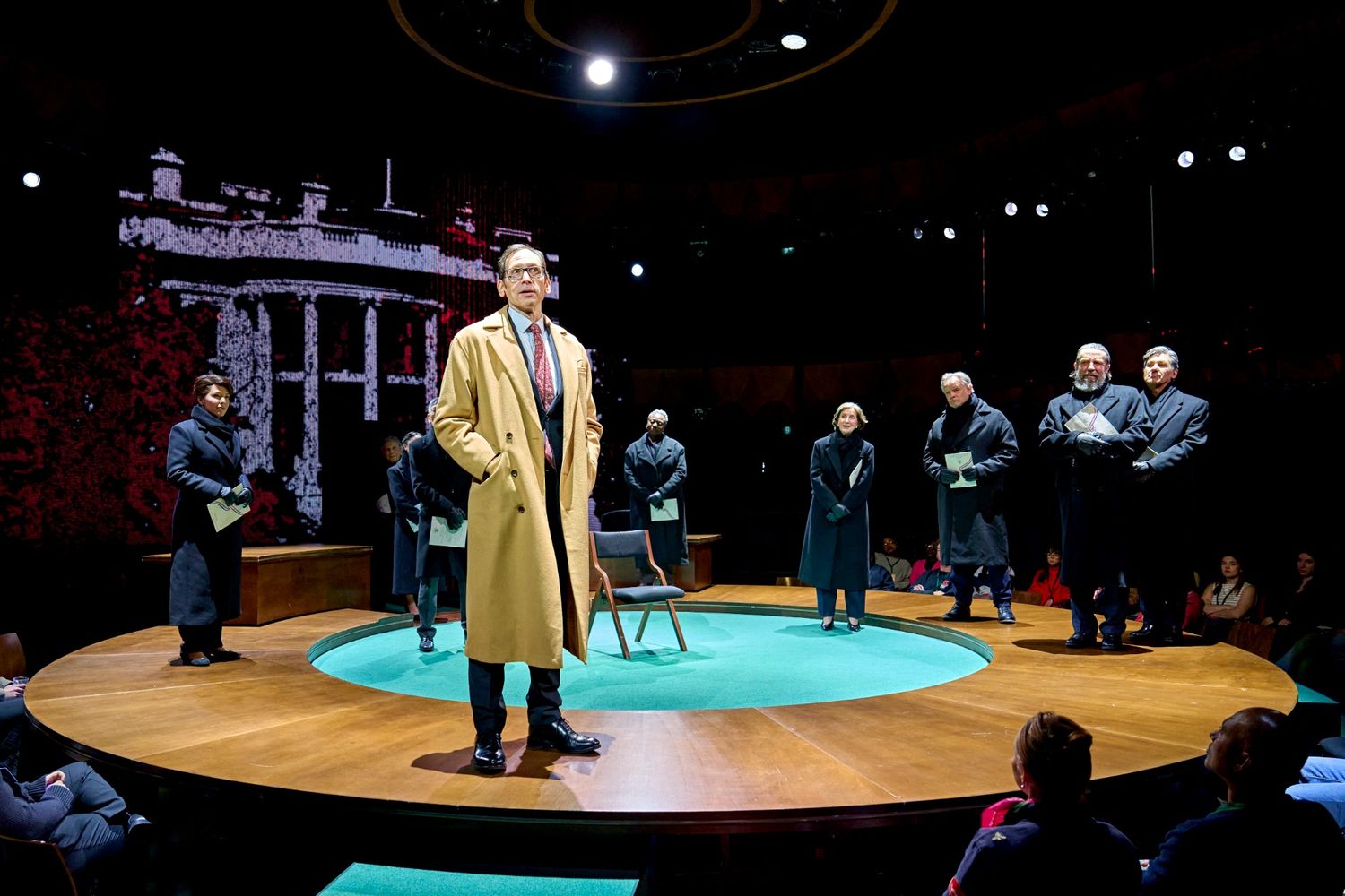 Stephen Kunken as Don Pearlman wearing a camel-coloured trench coat standing in front of the company wearing black trench coats.
